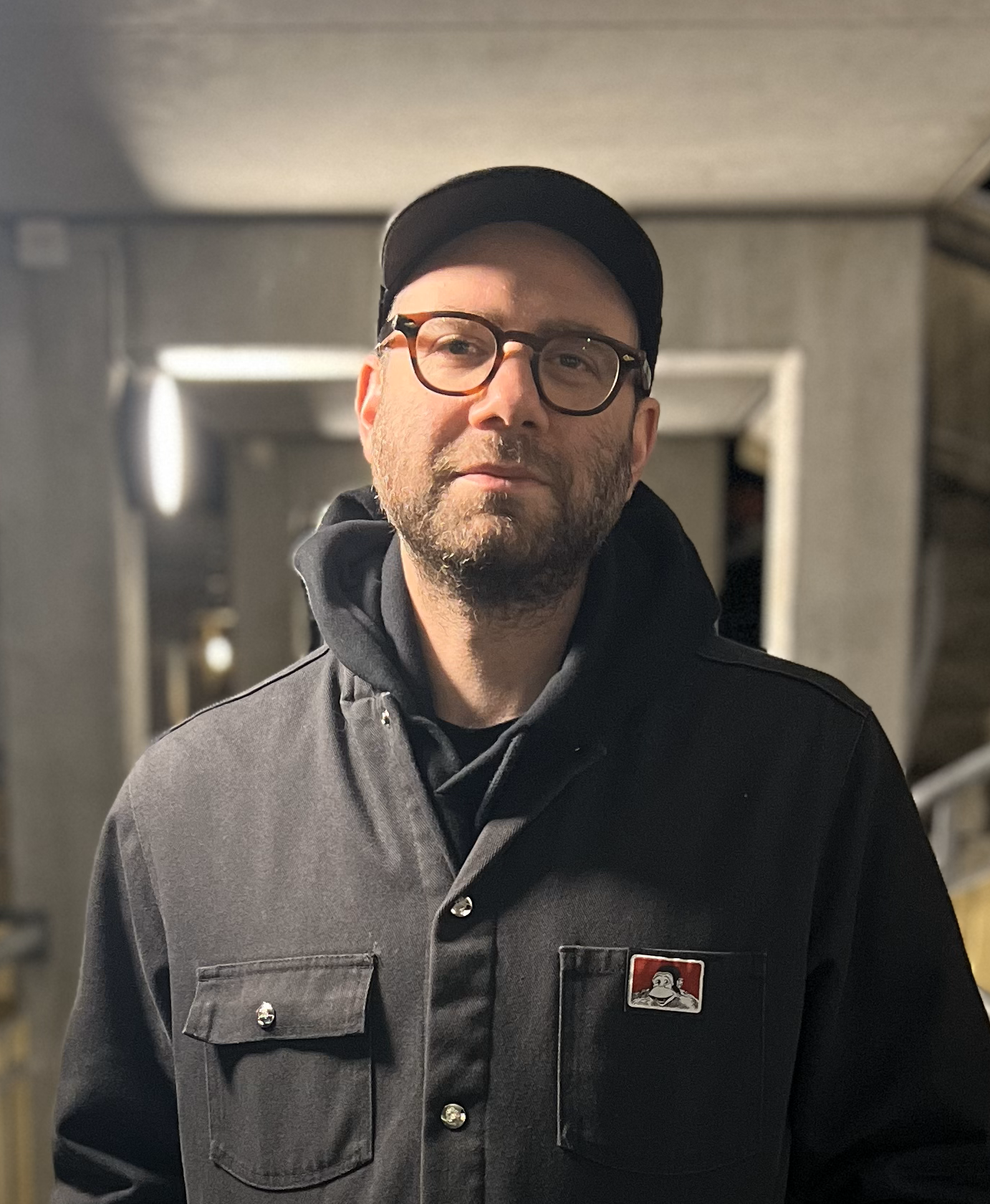 Man with dark blue jacket, hat and glasses, with shadow beard