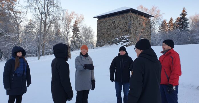 Kuusi ihmistä seisoo talvella lumisessa maisemassa. Ihmiset ovat ryhmänä kuvan etualalla ja keskustelevat toistensa kanssa. Taustalla mäen päällä näkyy kivinen torni ja puita.