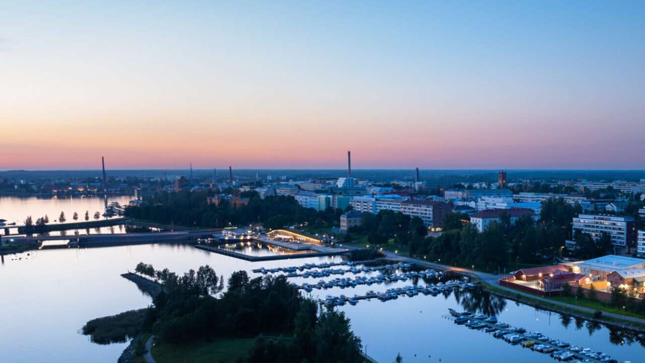 Aerial view of a marina and waterfront area in Vaasa during a summer evening, with boats docked in the calm water and city buildings illuminated against a pink and blue sky.