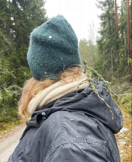 Katey's back to us as she looks into the forest. A moss-covered stick is attached to her scarf and hood.