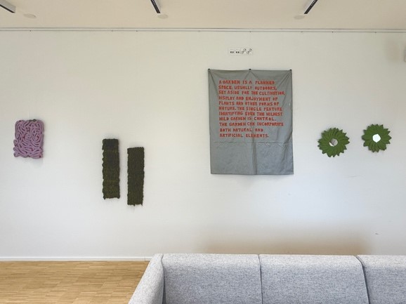 Installation picture of Jenni Österlund's exhibition, with a square artwork with text surrounded by circular textile pieces at the right, and two long rectangle textile pieces to the left and another smaller rectangular grey piece to the left.